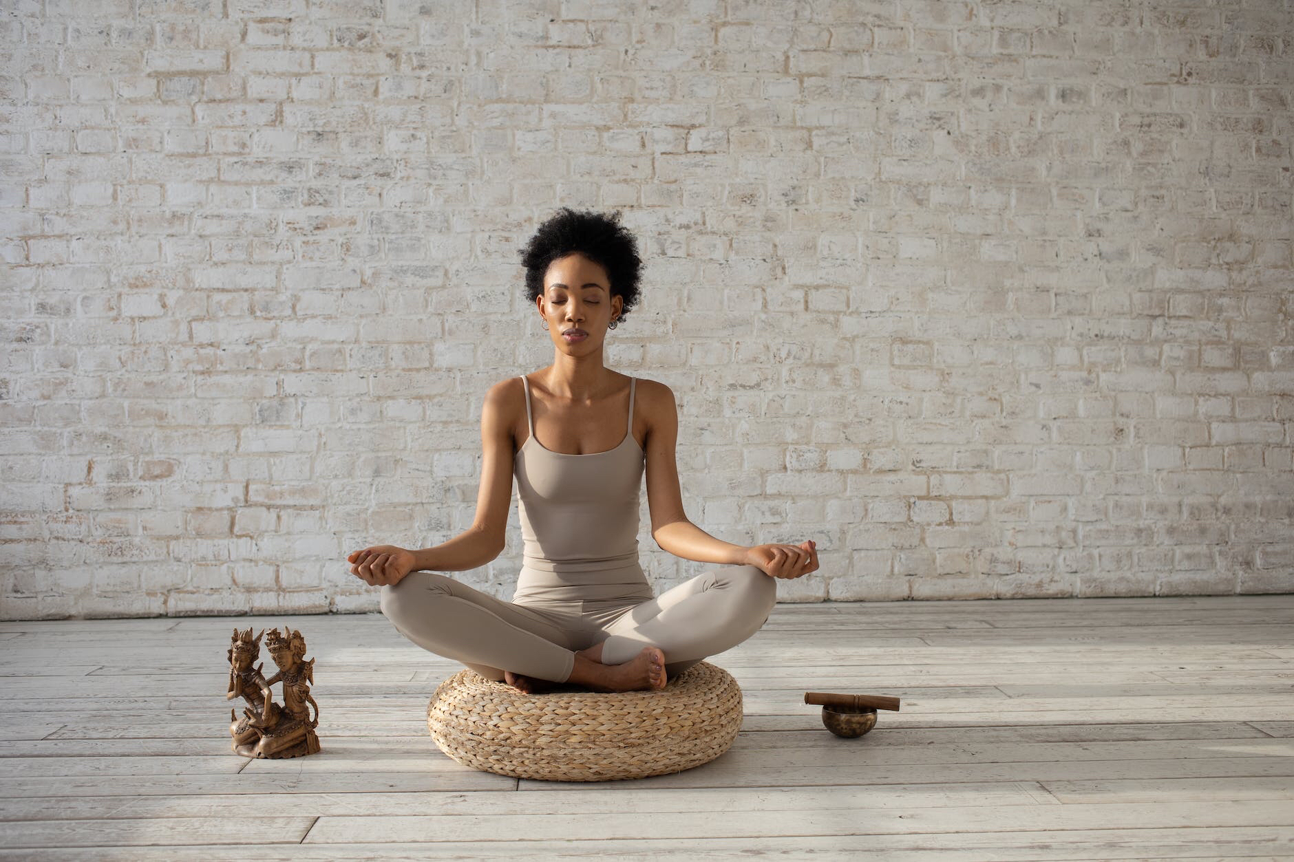 Woman meditating on a woven straw cushion in the lotus position in front of a whitewashed brick wall